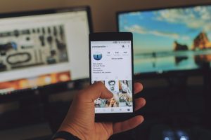 smartphon tenuto in mano e sullo sfondo due pc